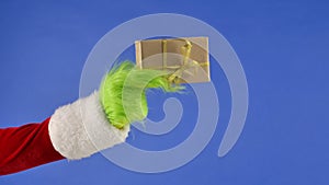 The Grinch's green haired hand holds a white gift box with a gold bow on an isolated blue background. Gift kidnapper