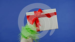 The Grinch's green haired hand holds a white gift box with a red bow on an isolated blue background. Gift kidnapper