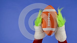 Grinch's green hairy hands holding out an American soccer on blue isolated background. Gift snatcher cosplay