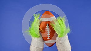 Grinch's green hairy hands holding out an American soccer on blue isolated background. Gift snatcher cosplay