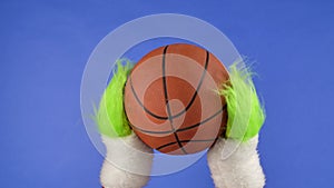 Grinch's green hairy hands holding out a basketball on blue isolated background. Gift snatcher cosplay. Christmas