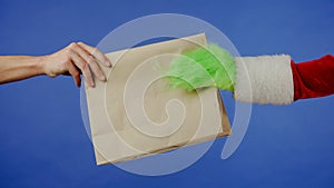 The Grinchs green haired hand reaches out and a mans hand takes a paper bag on an isolated blue background