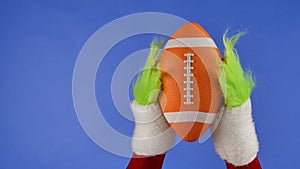 Grinchs green hairy hands holding an American soccer on blue isolated background. Gift snatcher cosplay. Christmas