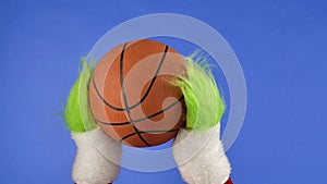 Grinchs green hairy hands holding a basketball on blue isolated background. Gift snatcher cosplay. Christmas and