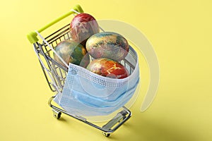 Grocery cart in protective mask with Easter eggs on yellow background, Easter time in quarantine, spring holyday during epidemy of