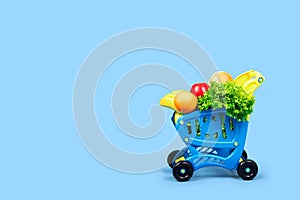 A grocery shopping cart full of salad, oranges, bananas, apples and bread. Close-up concept with place for text on a blue