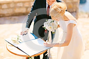 Groom in a black suit and bride in a white dress with a bouquet of flowers sign the marriage registration book