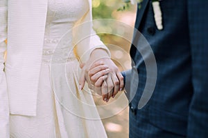 The groom in a blue suit and the bride in a wedding dress hold each others hands. Hands close up