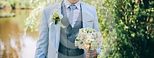 Groom in blue suit is standing outdoors. Handsome man is holding the wedding bouquet