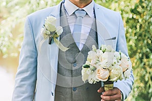 Groom in blue suit is standing outdoors. Handsome man is holding the wedding bouquet