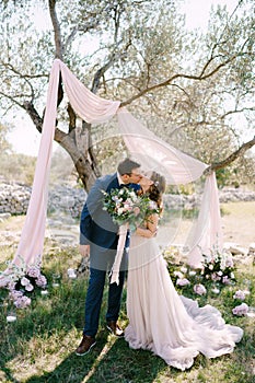 Groom and bride kiss under a tree in a green park