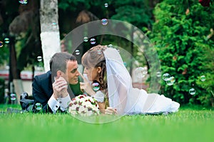 Groom and bride lie on a grass around soap bubbles
