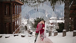 Groom comes to bride, hugs and kiss her in snowy mountain village at ski resort. Romantic wedding couple in knitted
