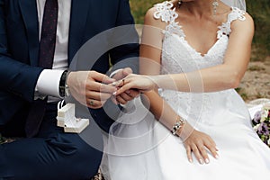 The groom in a dark blue suit puts a gold engagement ring on the finger of his bride
