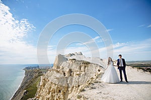 Groom in elegant suit and bride in white dress at weddingday on cliff with beautiful view of ocean