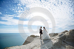 Groom in elegant suit and bride in white dress at weddingday on cliff with beautiful view of ocean
