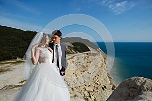 Groom in elegant suit and bride in white dress at weddingday on cliff with beautiful view of ocean