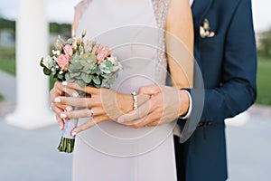 The groom embraces the bride, a large view of the hands. The bride holds a delicate bouquet of flowers