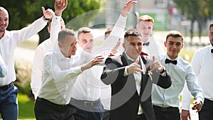 Groom flexing and having fun walking with groomsmen on wedding day. Happy classy man in black suit, bow tie and