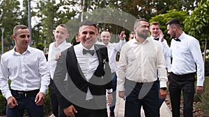 Groom flexing and having fun walking with groomsmen on wedding day. Happy classy man in black suit, bow tie and