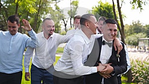 Groom flexing and having fun walking with groomsmen on wedding day. Happy classy man in black suit, bow tie and