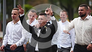 Groom flexing and having fun walking with groomsmen on wedding day. Happy classy man in black suit, bow tie and