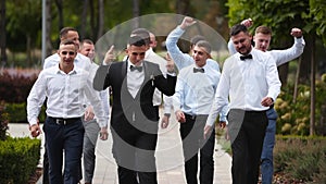 Groom flexing and having fun walking with groomsmen on wedding day. Happy classy man in black suit, bow tie and