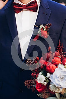 groom holding bouquet with red roses and succulents in the morning. rustic flowers. man in blue suit and red bow tie.