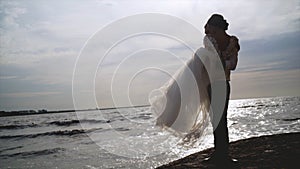 Groom holds bride in his arms on background of sea. Action. Groom picked up bride and turns her in sun on background of