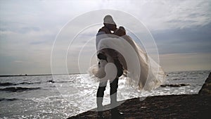 Groom holds bride in his arms on background of sea. Action. Groom picked up bride and turns her in sun on background of