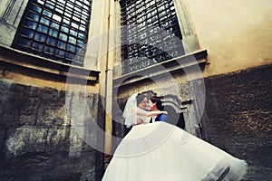 Groom holds bride on his arms standing under big windows