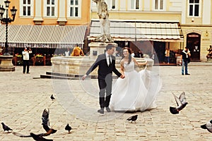 Groom holds bride`s hand while they run across a city square ti
