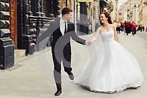 Groom holds bride`s hand tightly while they run along old buildi