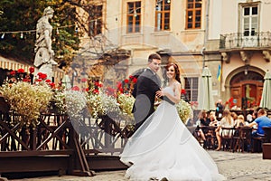 Groom holds bride`s waist posing behind a wooden street cafe