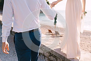 Groom holds the hand of bride with a bouquet, walking along the seashore