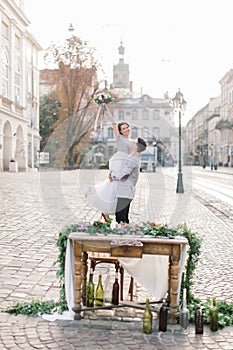 The groom holds his bride in his arms and stands against the background of old buildings in the city