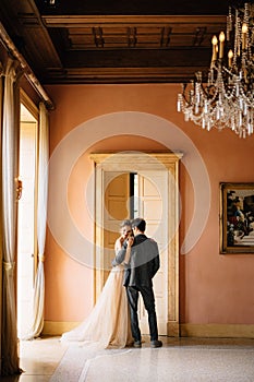 Groom hugs bride against the background of the high door to the room of the old villa. Lake Como