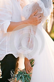 Groom hugs bride with a bouquet by the forearms. Cropped. Faceless. Side view