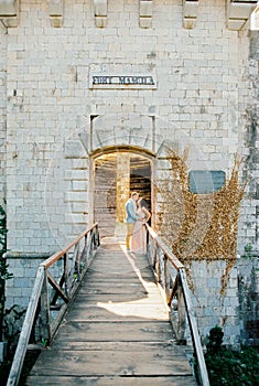 Groom hugs bride leaning against the railing of the wooden bridge to Mamula Fort