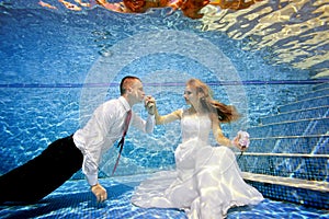 The groom kisses the bride`s hand, which is sitting underwater at the bottom of the pool. Portrait.