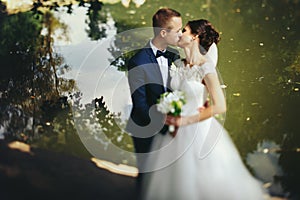 Groom kisses a bride standing on the lake shore