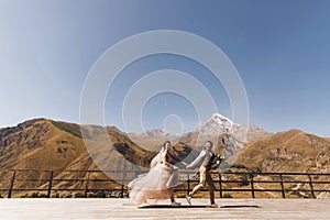 Groom in modern suit and bride in charming pink dress pose on the terasse with great mountain view in Georgia