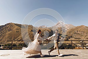 Groom in modern suit and bride in charming pink dress pose on the terasse with great mountain view in Georgia