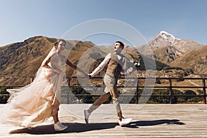 Groom in modern suit and bride in charming pink dress pose on the terasse with great mountain view in Georgia