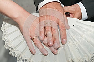 Groom`s hand with a gold ring on the bride`s hand also with a ring and both hands on a white fan.