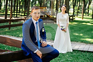 Groom is sitting on a wooden fence outdoor with beauty bride in blur on background