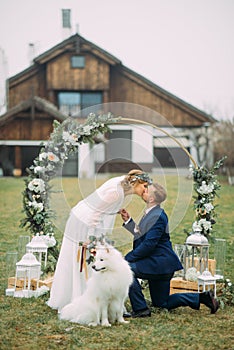 The groom stands on one knee and kisses his bride next to dog and a wedding arch