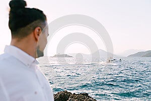 Groom stands on the rocks and looks at the motor yacht sailing on the sea