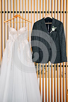 Groom suit and bride dress hang on hangers on a wooden plank wall in a hotel room