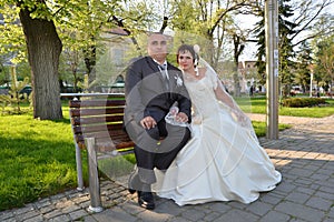 The groom in a suit and the bride in a white dress are photographed on a bench in a city park on a background of fresh green grass